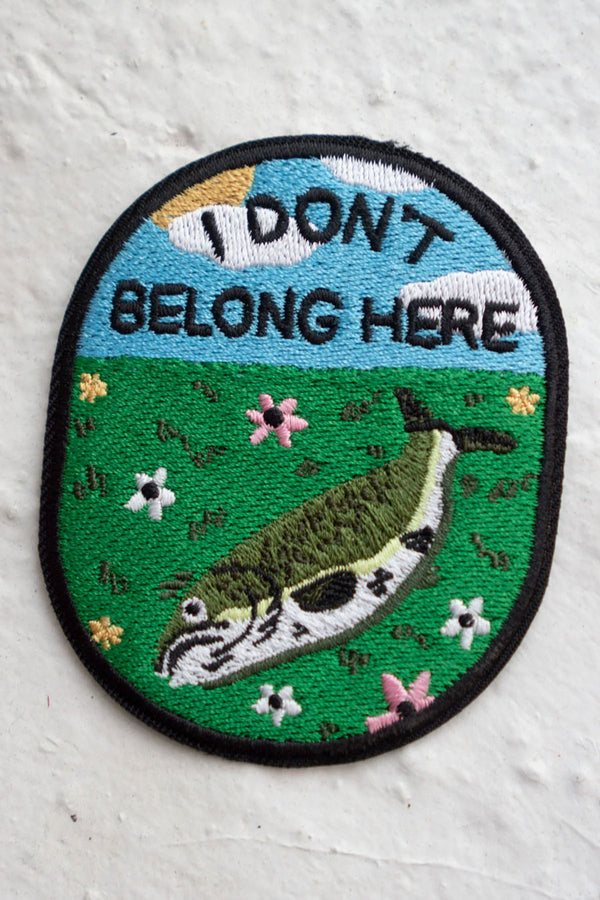 Oval patch that features a fish that is laying on green grass surrounded buy small flowers underneath a blue sky. In the sky, in black text, the patch says I don't belong here. White background.