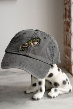 Gray denim baseball hat featuring a green and pink jumping frog. the hat is sitting ontop of a small dog statue. 
