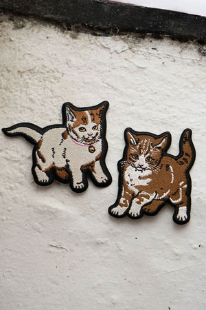 Embroidered pair of kitten patches. The kittens are tan and white, one with a pink collar. White concrete background.