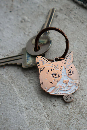 Soft enamel keychain with copper hardware. The keychain features a portrait of a cat with scowling face. The cats collar says Lil' Shit. Concrete background.