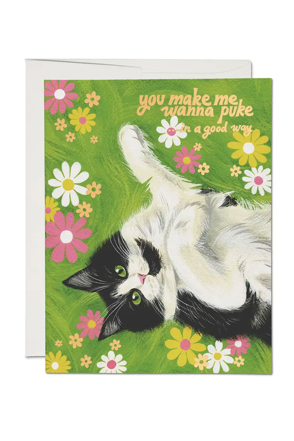 Greeting card of an illustration of a black and white cat laying in the grass surrounded by pink, white, and yellow flowers. The card says You Make Me Wanna Puke. In a Good Way. White background.