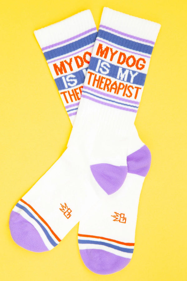 Mid calf athletic socks with a purple heel and toe. and Blue, purple and green stripes across the tops. Blue and red stripes across the toes. The socks say My Dog is my Therapist in red lettering. Yellow background.