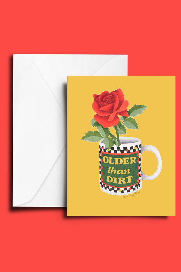 Yellow greeting card with white envelope against a red background. The card features an illustration of a coffee mug with a red rose in it. The mug says Older than Dirt. 