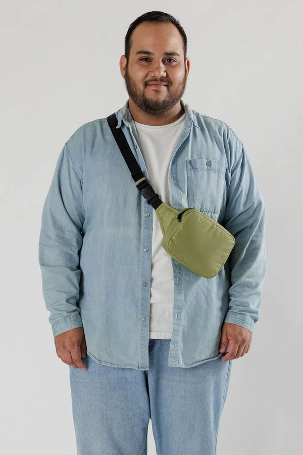 A man wearing a denim button up and white tshirt standing in front of a white backdrop. He is wearing a puffy pistachio green fanny pack with black straps across his body.
