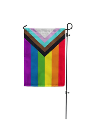 Progress Pride flag hanging from a garden pole against a white backdrop. The Progress Pride Flag includes a black, brown, blue, pink and white stripe layered on top of the rainbow colors.