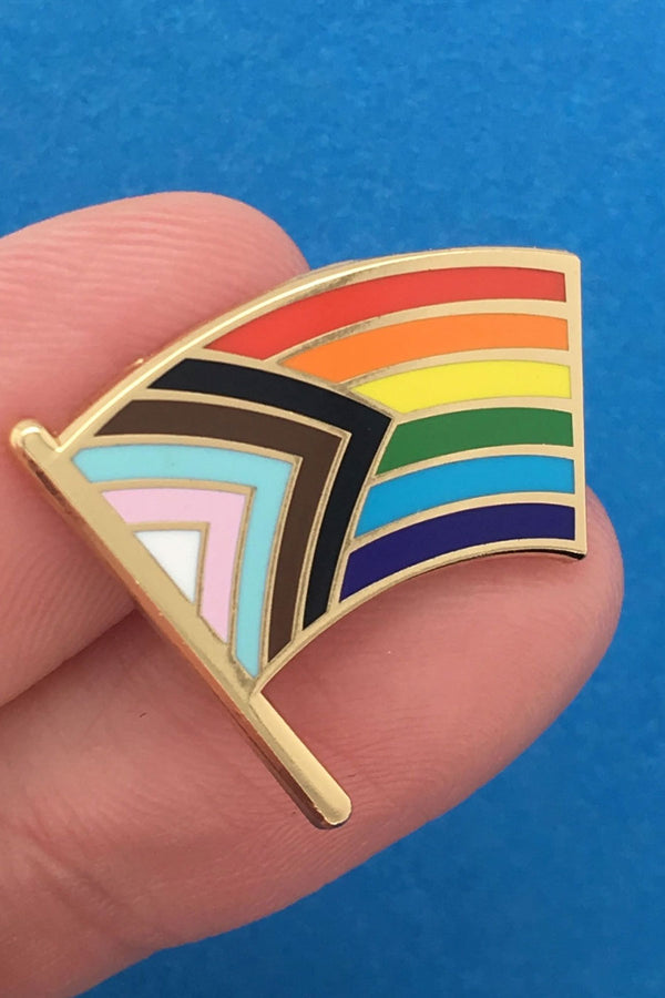 Someone holding an enamel pin of the progress pride flag.