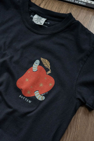 Black tshirt laying on a wooden table. The tshirt features an illustration of a red apple with gold stars and a green worm sticking out of it. The worm is frowning and below the apple in green text it says ROTTEN. 