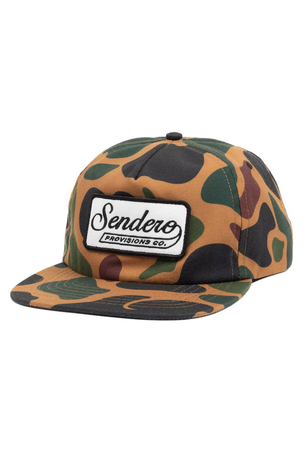 camouflage pattern baseball cap. The front of the cap has a white patch with a black border. The patch says Sendero Provisions Co.
