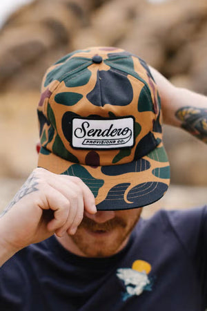 Man wearing camouflage pattern baseball cap. The front of the cap has a white patch with a black border. The patch says Sendero Provisions Co.  