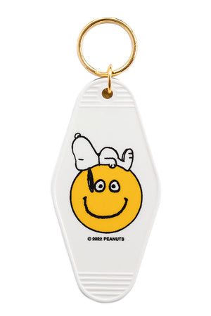Vintage motel style keychain in white. The keychain features Snoopy laying on top of a yellow smiley face. Gold hardware. White background.