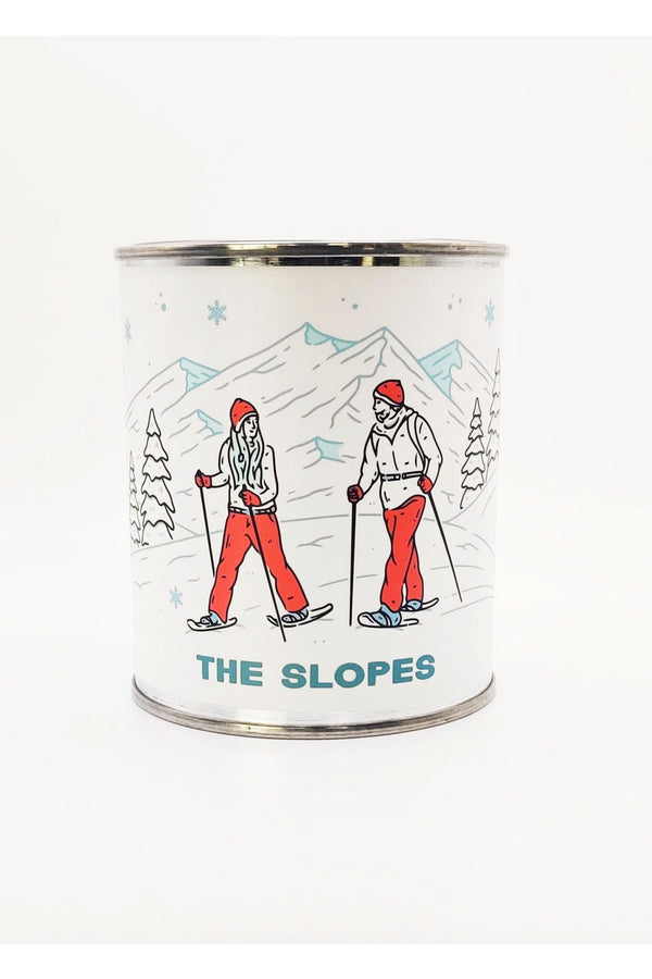 Tin Candle with illustrated label of two people skiing against a snowy mountain backdrop. The candle is called The Slopes.