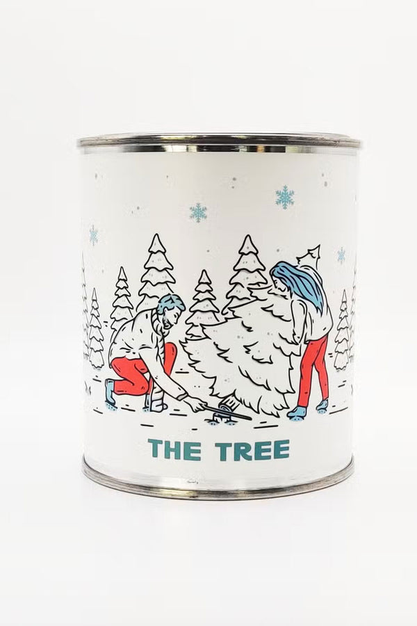 Candle in a metal tin container. The label is white and features an illustration of two people cutting down a tree in the snow. The candle is called The Tree. 