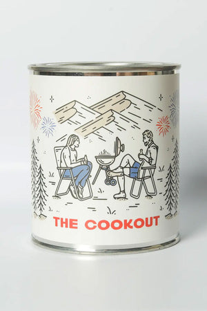 Tin candle called The Cookout. Label features a couple sitting in camping chairs around a bbq grill with fireworks in the sky and a mountain range in the background.