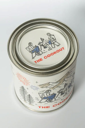 Tin candle called The Cookout. Label features a couple sitting in camping chairs around a bbq grill with fireworks in the sky and a mountain range in the background.