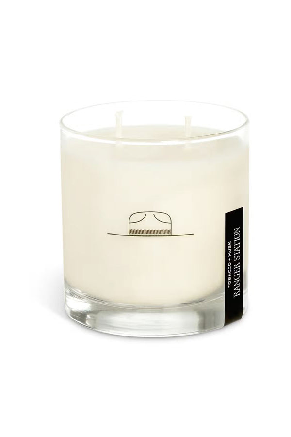 Soy candle in clear glass container with a Park Ranger hat logo on the front. the candle has a small black label that says Ranger Station. Scent is Tobacco and Musk. White background.