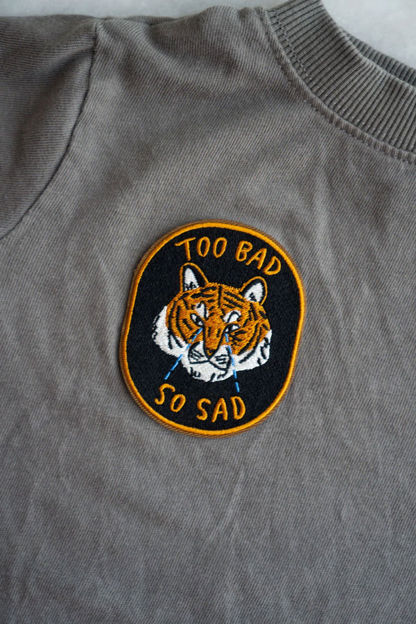 A Grey tshirt featuring a black oval patch with orange border. The patch features a crying orange tiger with the words "Too Bad So Sad" in orange text.