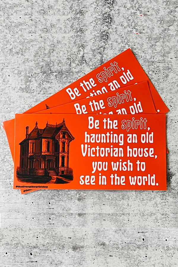 Red bumper sticker of a black victorian house drawing. Next to the house in white spooky lettering it says Be the spirit, haunting and old victorian house, you wish to see in the world.