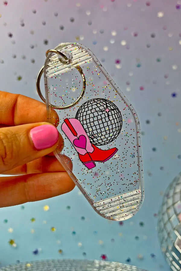 Person holding Clear glitter vintage motel style keychain with a disco ball and red and pink cowboy boots with a red heart on them. Blue glitter background.