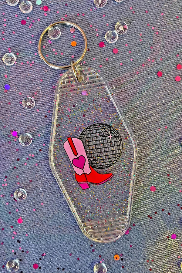 Clear glitter vintage motel style keychain with a disco ball and red and pink cowboy boots with a red heart on them. Blue denim with pink confetti background.