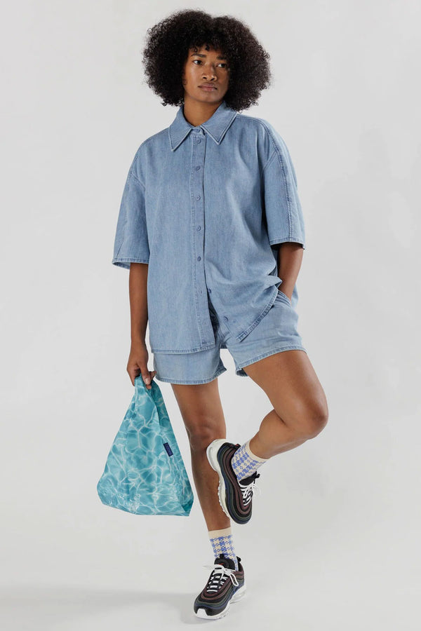 A person wearing a denim top and denim shorts holding a Small reusable shopping back that features a blue reflective water pattern from a pool. White background.