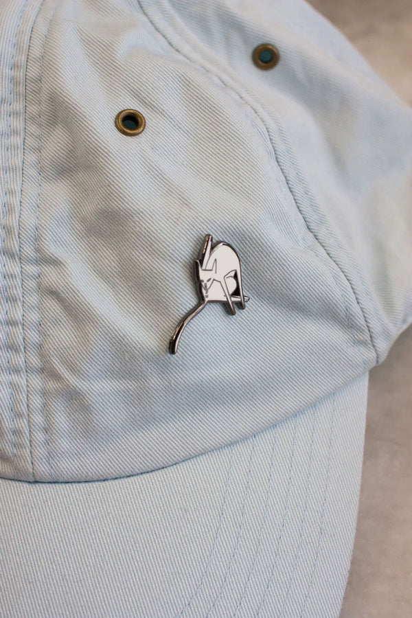 Enamel pin of a white cat bathing itself pinned to a light denim hat.
