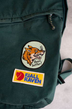 Close up view of a green backpack featuring a patch of Mint green oval patch with black border. The patch features a crying orange wolf that is crying. Below that the patch says Been Better in black text.