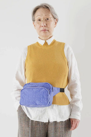 A person standing in front of a white wall wearing a light blue fanny pack with black nylon straps across their waist.