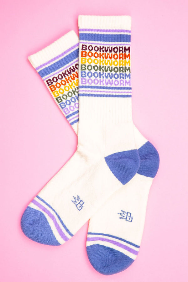 One pair of off white mid calf athletic sock with a blue heel and toe. Across the toes and the top are muted purple and blue stripes. The socks say Bookworm six times. Each one with a different mutes color assigned to it. Top to bottom is Maroon, orange, yellow, green, blue, and purple. Pink background.