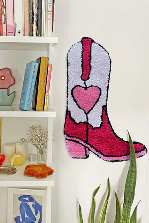Pink western style cowboy boot rug with a heart in the center. The rug is hanging on a white wall next to a bookshelf.