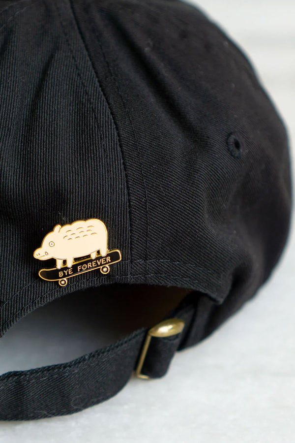 Enamel pin on the back of a black baseball cap. The pink is of a light peach boar with a tear falling from its eye. It is riding a skateboard that says Bye Forever.