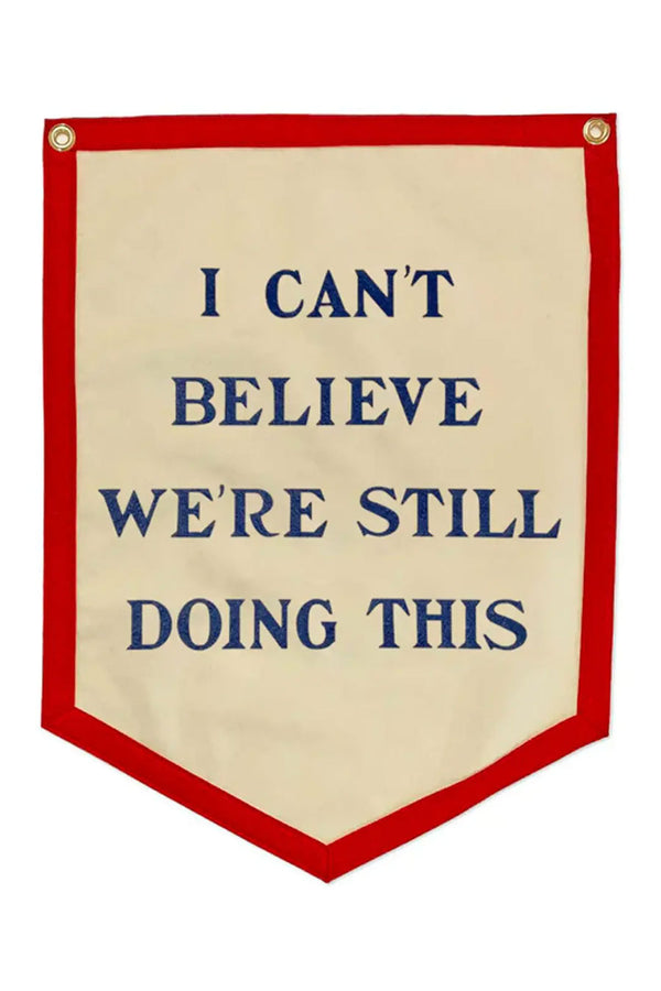 Ivory Camp flag that comes to a point with a red border and two gold grommets at the tope. The flag says "I Can't Believe We're Still Doing This" in blue lettering. White background.