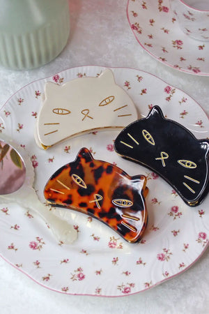 Photograph of three hair claw clips in the shape of a cats face. There is a white, black, and tortoise version of the clips laid out on a floral plate.