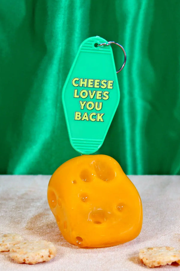 Green vintage motel style keychain. The keychain is sitting on top of a small piece of cheese in front of a green satin background. The keychain says Cheese Loves You Bakc in yellow lettering.