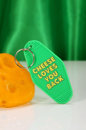 Green vintage motel style keychain. The keychain is leaning against a small piece of cheese in front of a green satin background. The keychain says Cheese Loves You Bakc in yellow lettering.