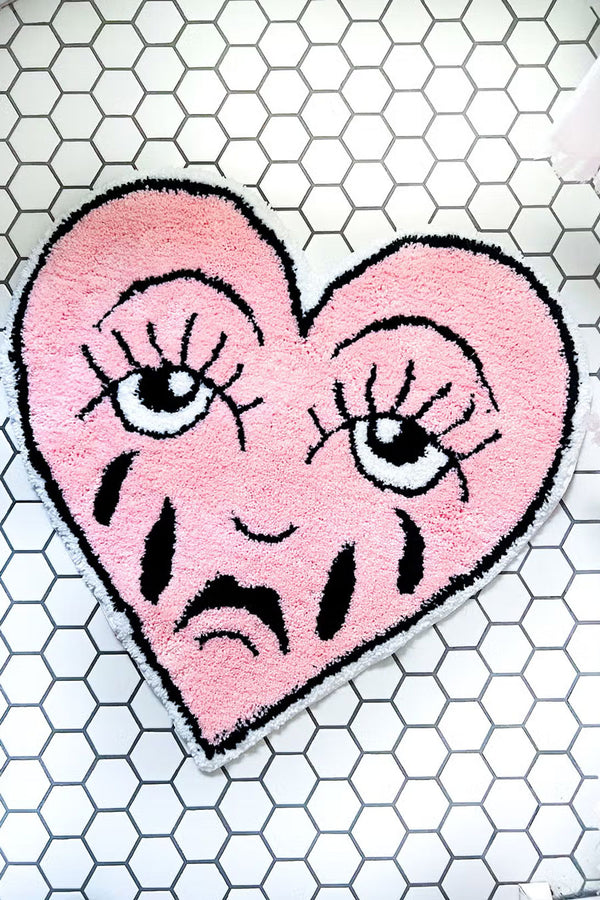 Heart shaped floor rug that features a crying face in traditional tattoo style. White tile floor background.