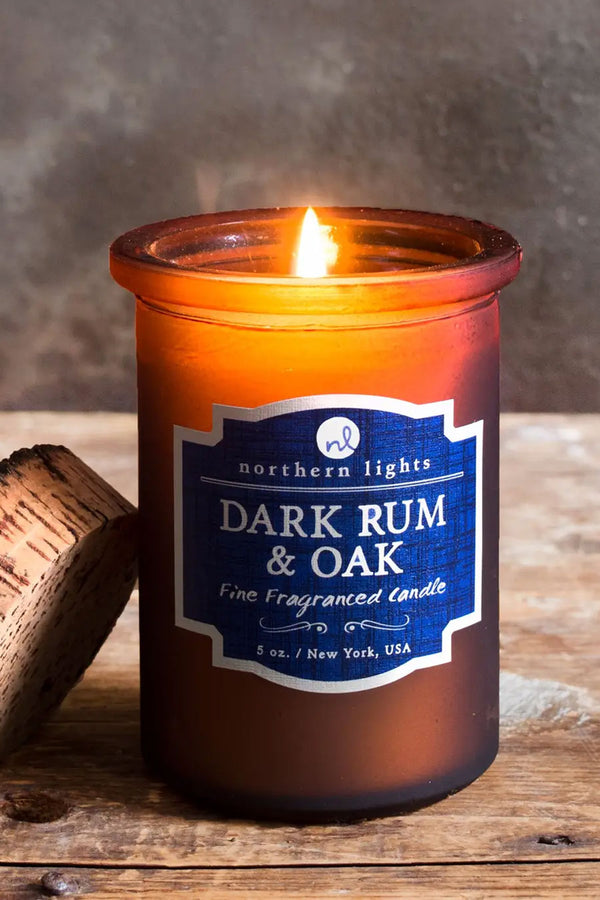 Frosted amber glass candle jar with a raw edge cork top. Candle has a blue label that says Dark Rum and Oak. Natural wood and stone backdrop.