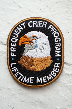 Oval shaped embroidered patch featuring a crying Eagle surrounded by text that says Frequent Crier Program. Lifetime Member. White concrete background.