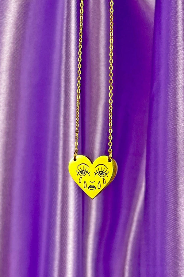 Gold heart necklace with gold chain. The heart features a crying face. Purple satin background.