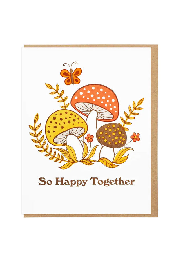 White greeting card with illustration of three mushrooms surrounded by flowers and grass with a butterfly overhead. Below the illustration the card says So Happy Together. Kraft brown envelope. White background.