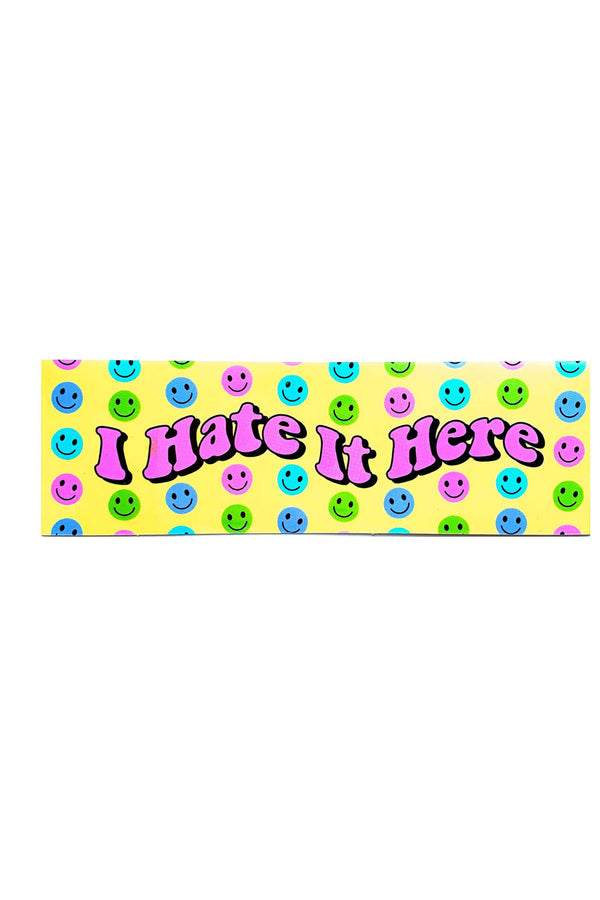 Yellow vinyl sticker that says I Hate It Here in pink bubble letters. Smiley faces in blue, aqua, green, and pink surround the lettering.