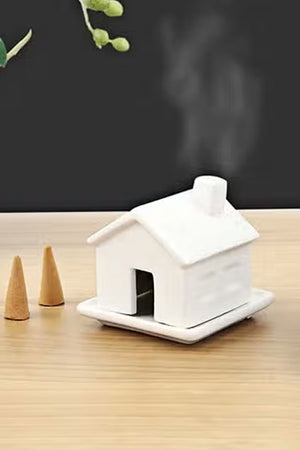 Small white house shaped incense burner on a wooden tabletop next to a couple of incense cones.