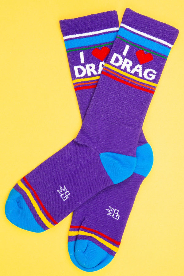 One pair of purple mid calf athletic socks with a blue heel and toe. Across the toes there is yellow and red stirpes. Across the top there is White, blue, green, yellow, pink, and red stripes. The socks say I Heart Drag in white lettering. Yellow background.