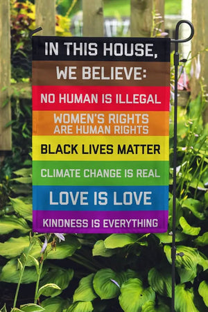 Photo of a garden with a garden flag with the following text on a rainbow field: IN THIS HOUSE, WE BELIEVE: NO HUMAN IS ILLEGAL WOMEN'S RIGHTS ARE HUMAN RIGHTS BLACK LIVES MATTER CLIMATE CHANGE IS REAL LOVE IS LOVE KINDNESS IS EVERYTHING.