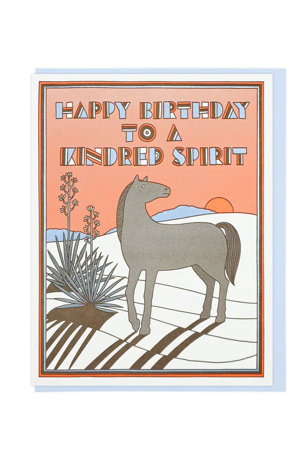Greeting card featuring a horse standing next to a desert plant in front of a sunset. Above the horse the card says Happy Birthday to a Kindred Spirit. Blue envelope. White background.