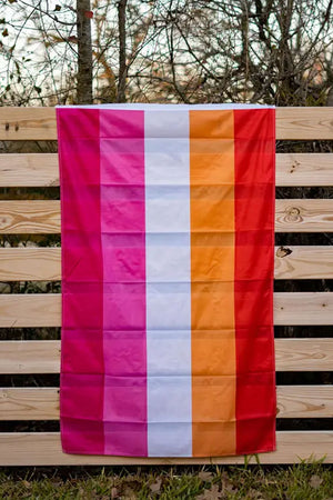 Photograph of the Lesbian Pride Flag hanging from a wooden fence. The flag consists of red, orange, white, pink, dark pink stripes.
