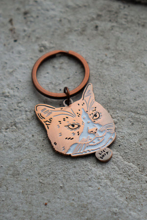 Soft enamel keychain with copper hardware. The keychain features a portrait of a cat with scowling face. The cats collar says Lil' Shit.