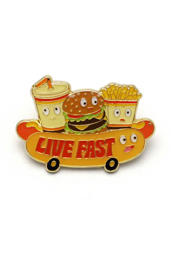 Enamel pin of a hot dog on wheels. On top of the hot dog is a cheeseburger, a milkshake, and Fries. Each item has a smiley face. On the bun of the hot dog the pin says Live Fast in red lettering. White background.