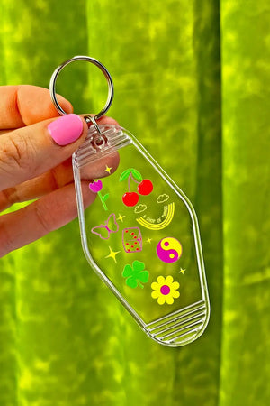 Person holding clear vintage style motel keychain. Keychain features mini illustrations of dice, four leaf clover, butterfly, pink tulip, yink yang, daisy, and cherries. Green background.