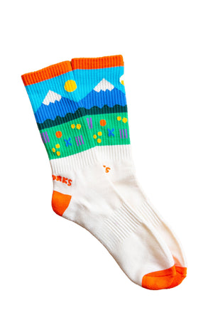 Crew socks featuring a mountain range in front of a green pasture.  The socks have orange heels and toes and a orange band across the top.