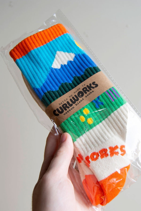 Crew socks featuring a mountain range in front of a green pasture. The socks have orange heels and toes and a orange band across the top.